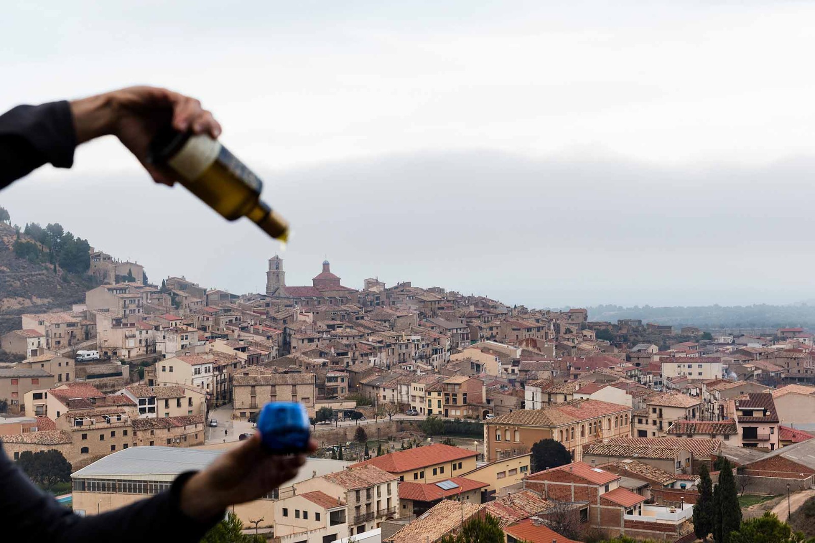 Imagen de una botella de aceite de Mas dels Castellans que está siendo servida en una copa, y al fondo se ve una vista del pueblo de Calaceite